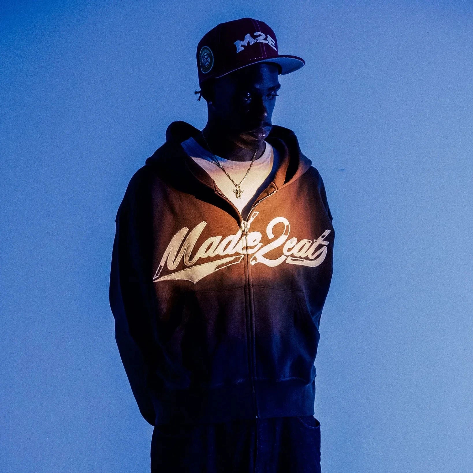 man in front of a blue backdrop wearing the brown M2E rope stitched hoodie in streetwear fashion.