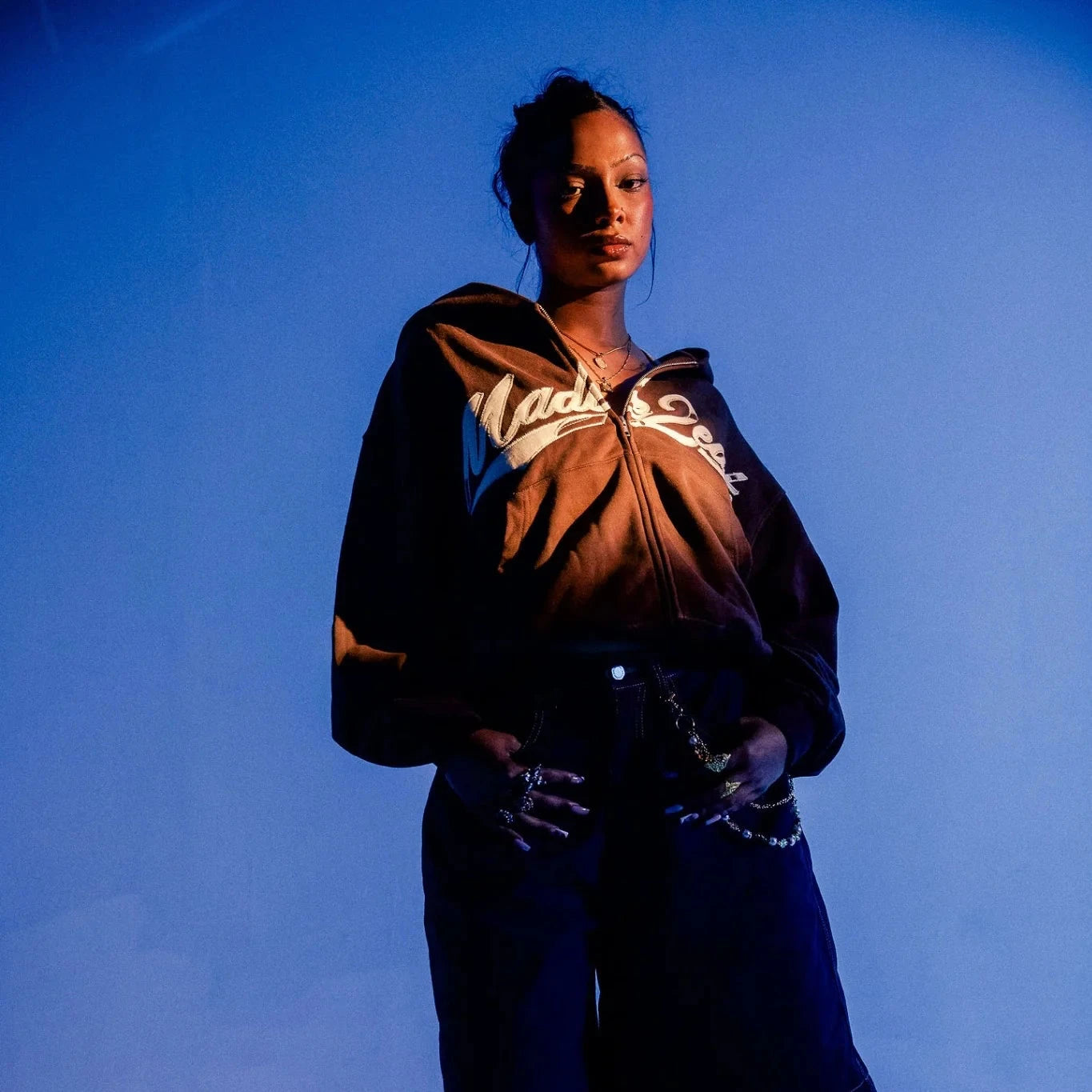 woman in front of a blue backdrop wearing the brown M2E rope stitched hoodie in streetwear fashion.