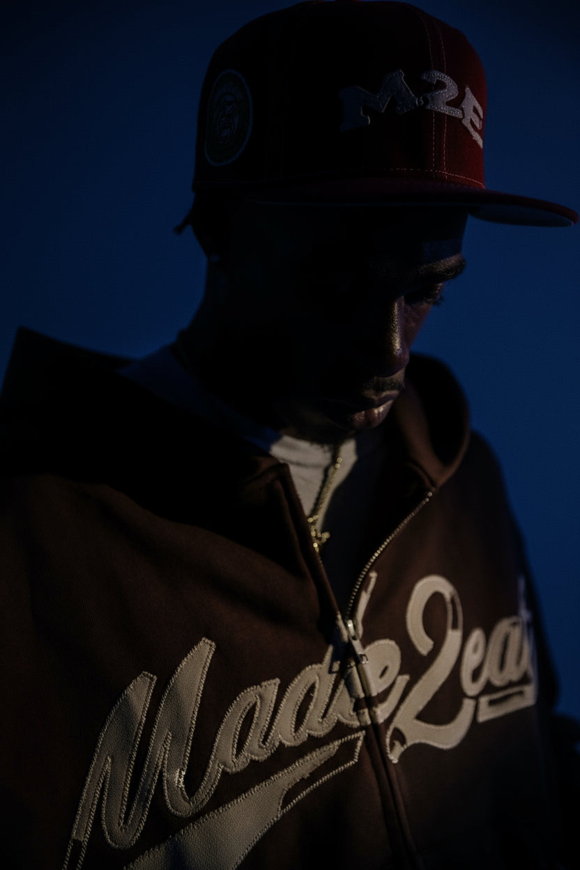 Person wearing a brown jacket with 'M2E' branding and a red cap with a logo against a blue background