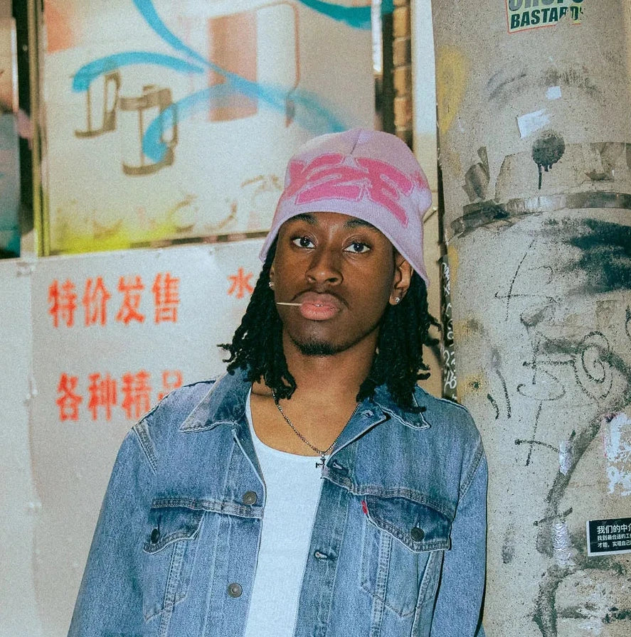 Person wearing a denim jacket and pink M2E beanie standing next to a graffiti-covered wall.