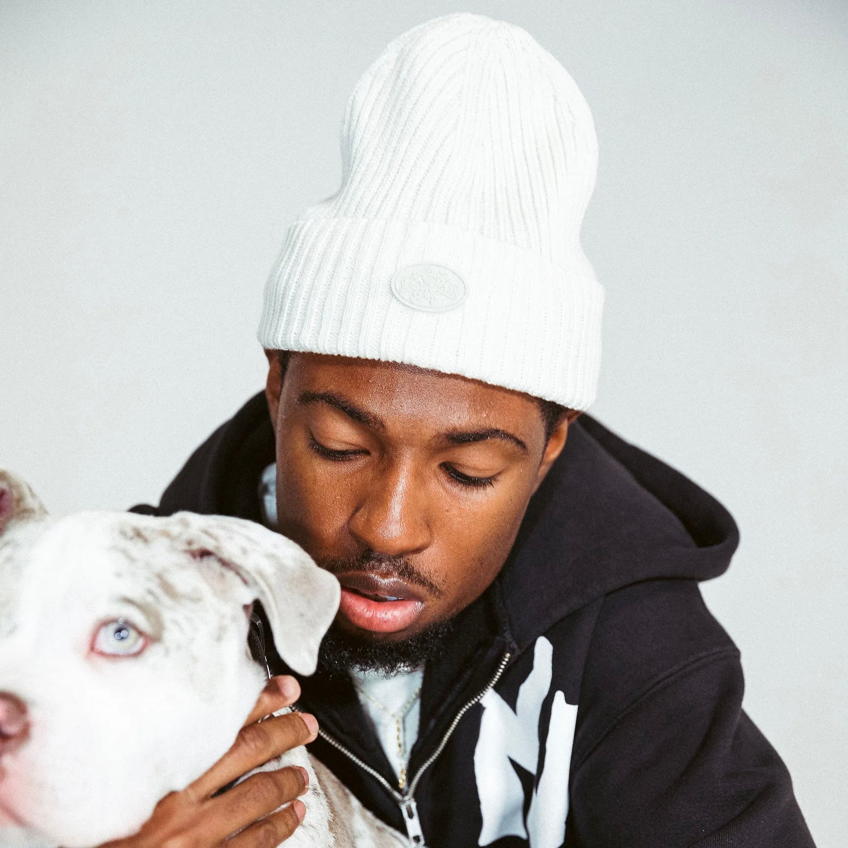 Man wearing a cream M2E skully and black hoodie with a dog on a light gray background