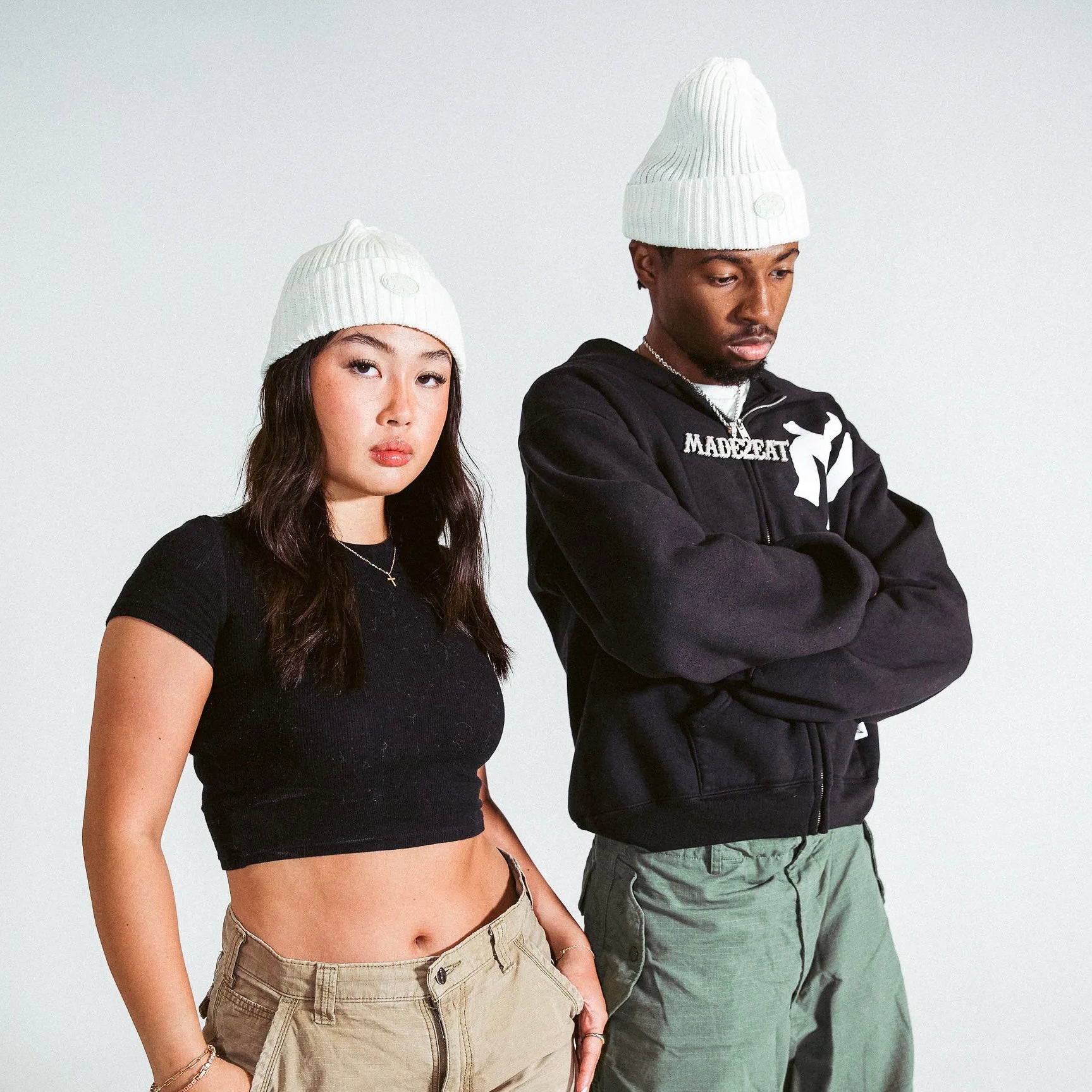 Two people standing wearing cream M2E skully beanies in front of a grey backdrop