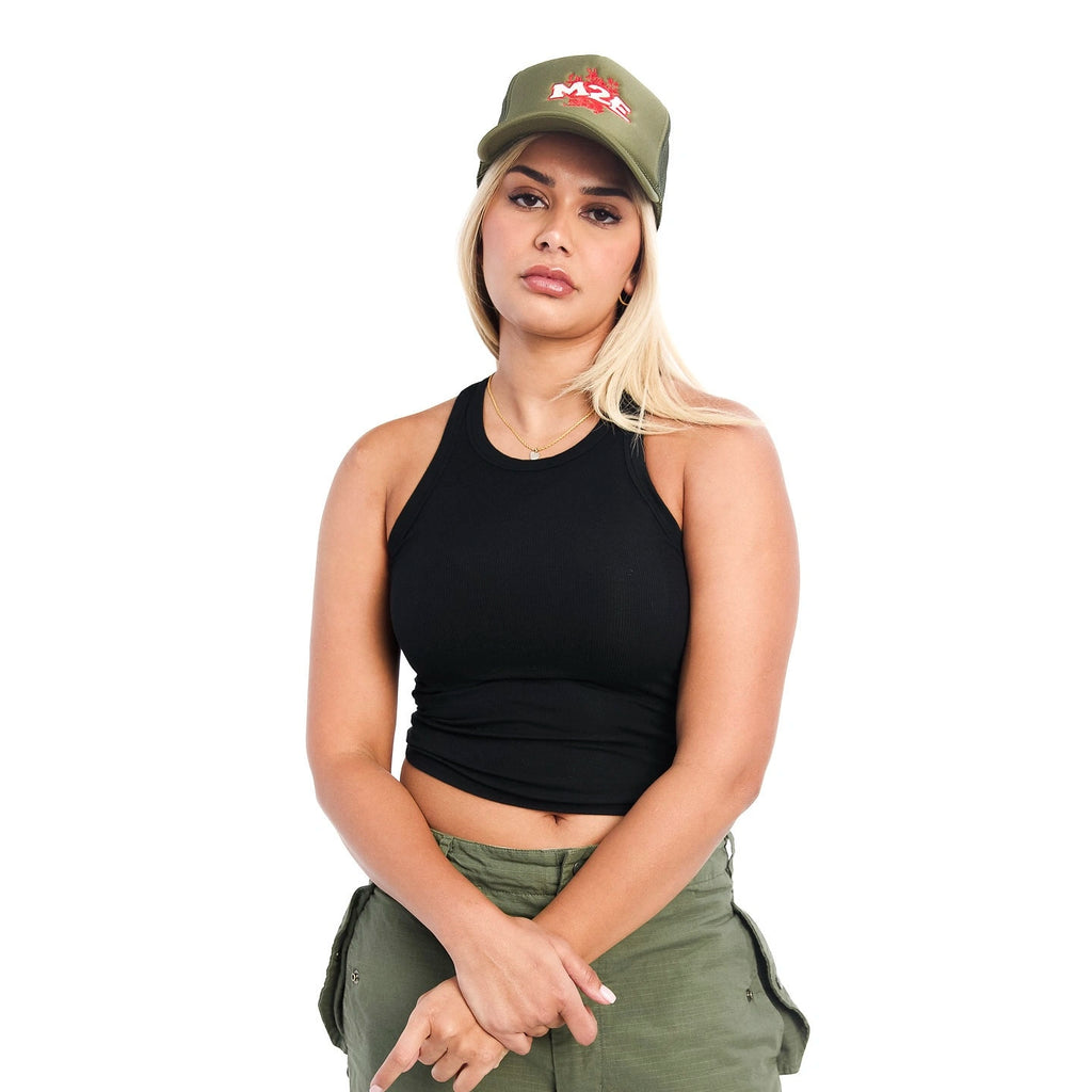 Woman wearing a black tank top, green M2E trucker cap, and green pants on a white background